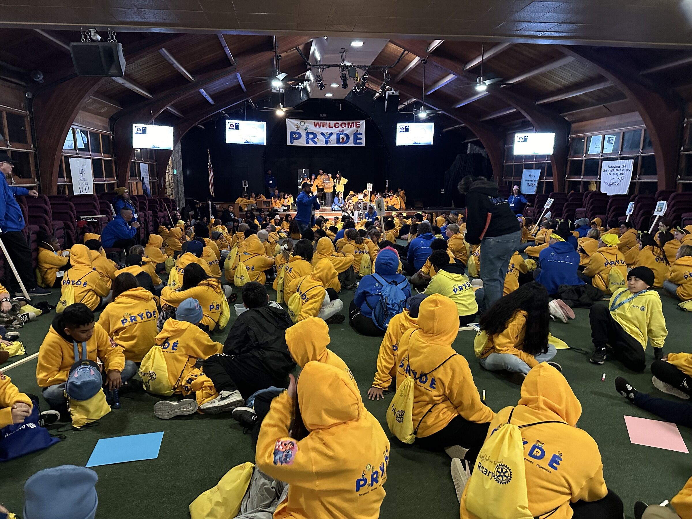 Campers in yellow PRYDE sweatshirts seated together in the main hall beneath the Welcome PRYDE banner