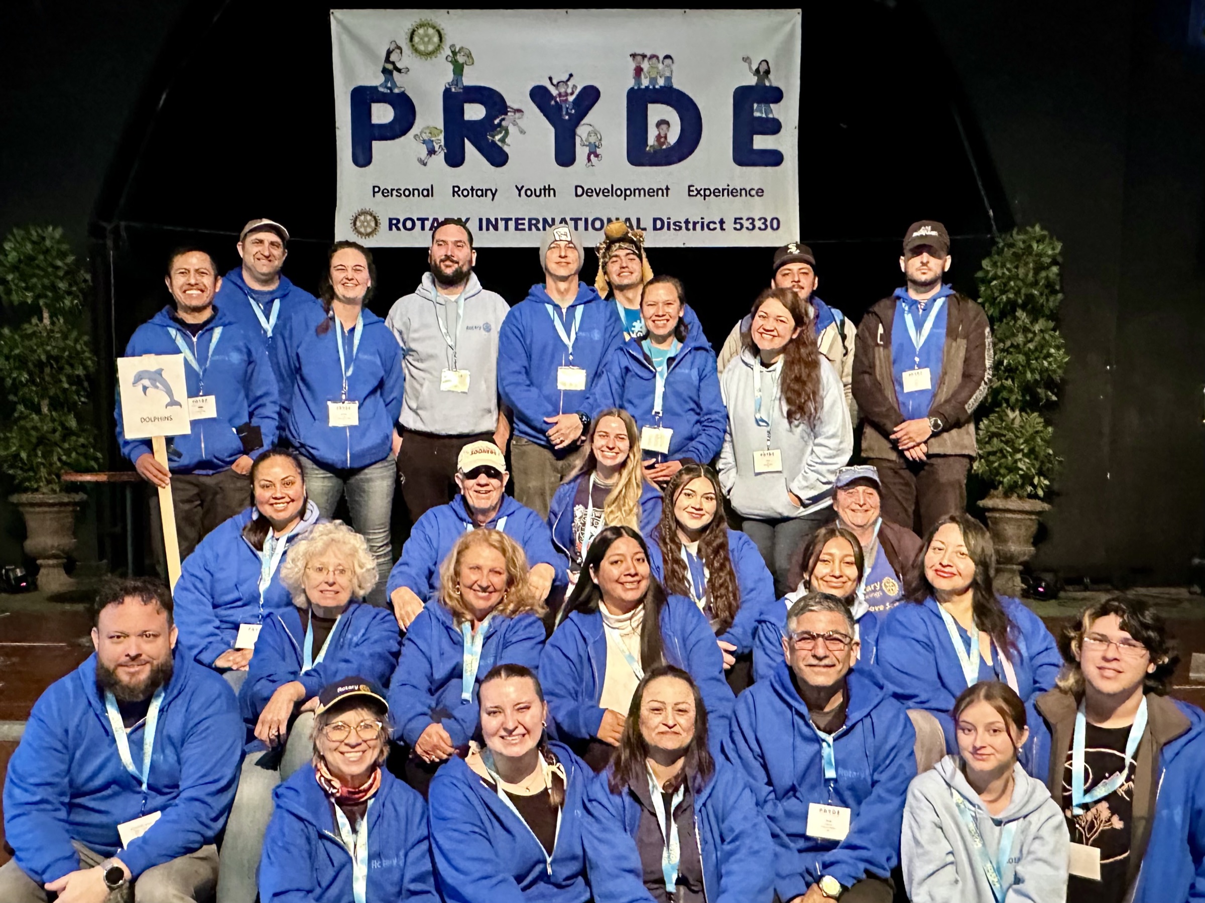 Rotarian volunteers in blue PRYDE jackets gathered beneath the PRYDE banner