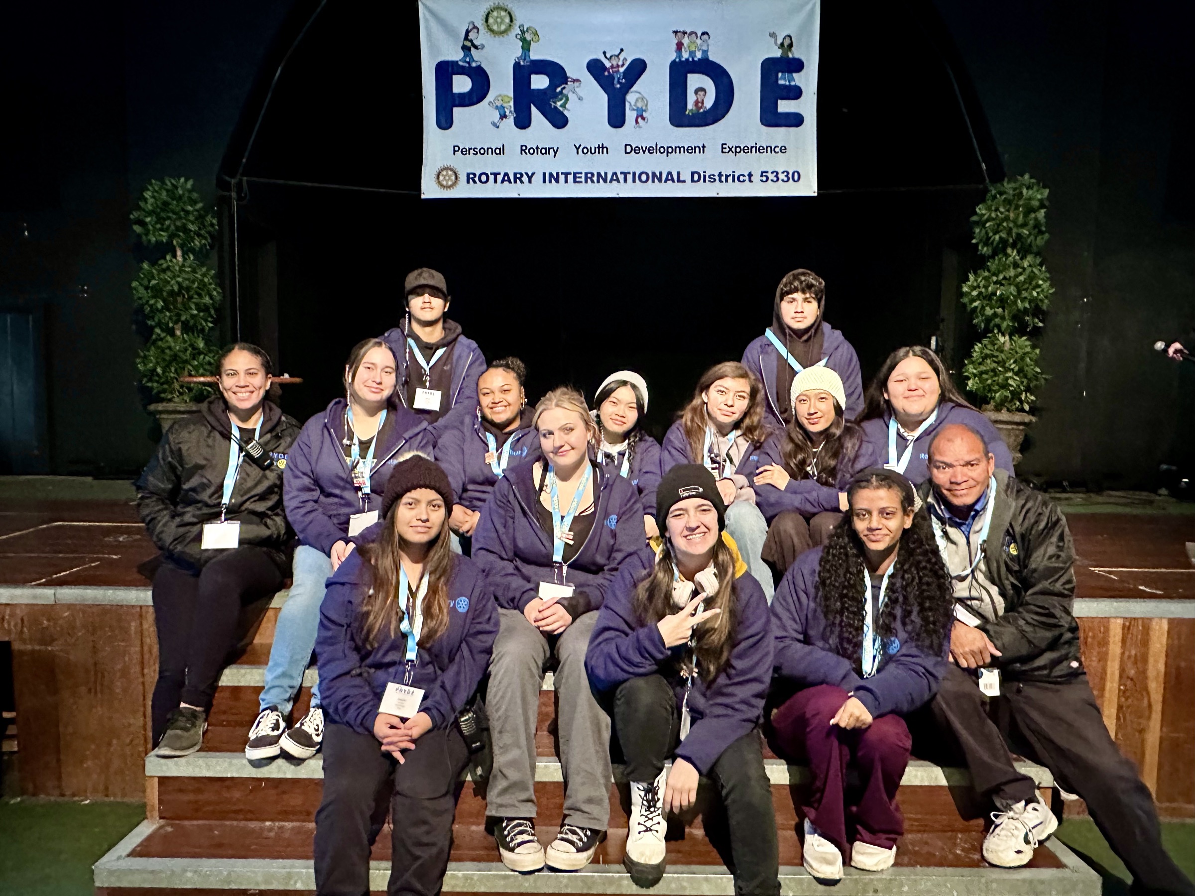 PRYDE Guides seated together on the stage steps beneath the PRYDE banner